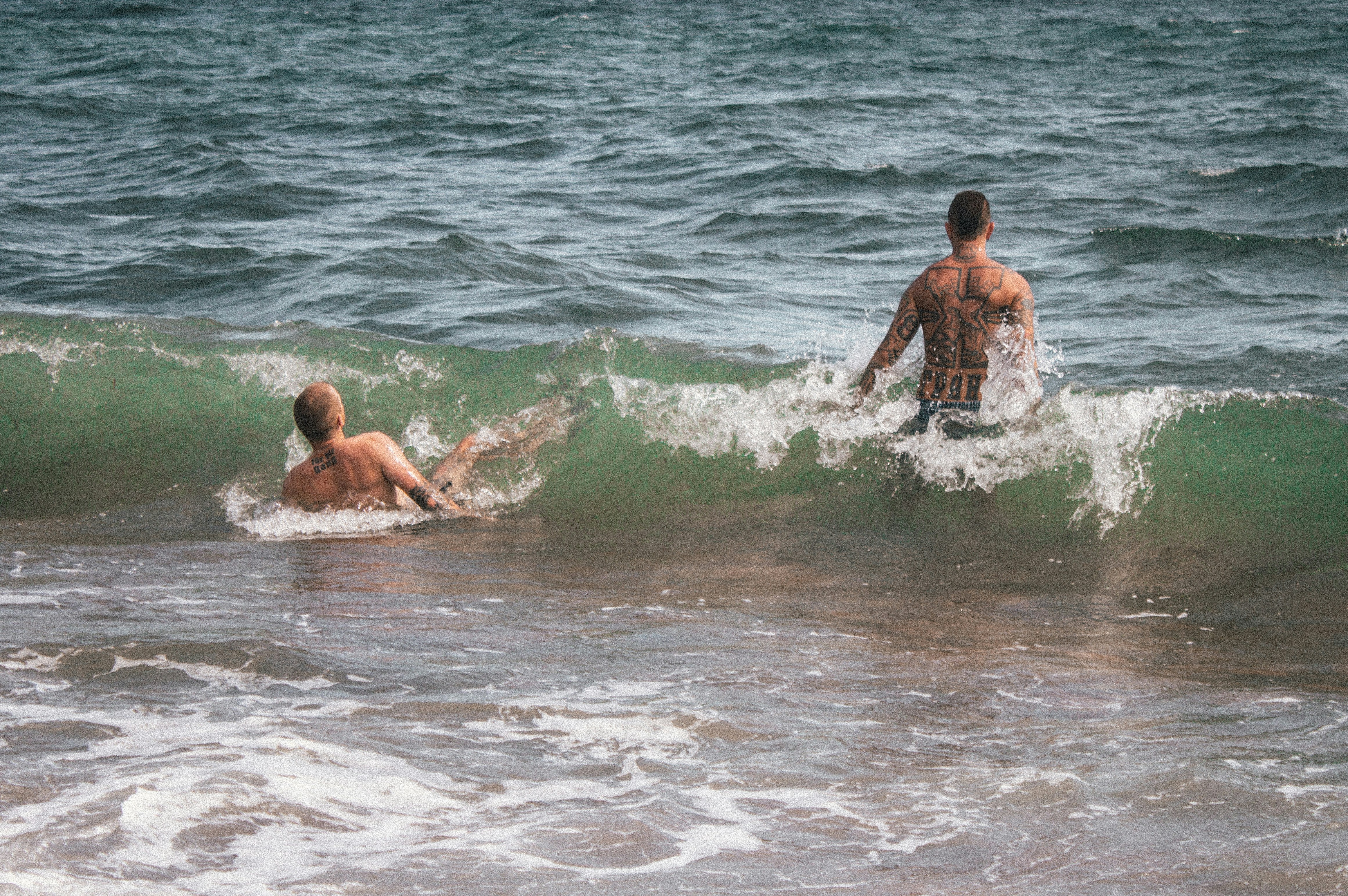 Two individuals with tattoos are enjoying the ocean waves. One person is standing, facing the water, while the other is partially submerged and lying back against the incoming wave. The water appears powerful and the setting is a beach with visible surf.