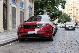 A red Range Rover is parked on a cobblestone street, surrounded by classical architecture and other vehicles. The scene seems to be in an urban area with a tree-lined street. Buildings with ornate designs are visible, contributing a historical and sophisticated aspect to the environment.