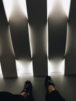 The image presents a pair of black shoes worn by an individual seated indoors. The setting includes vertical, white, illuminated panels casting a soft, ambient light across the floor. The overall feel is minimalistic and modern, with strong angular lines and contrasting light and shadow.