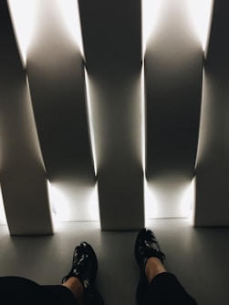 The image presents a pair of black shoes worn by an individual seated indoors. The setting includes vertical, white, illuminated panels casting a soft, ambient light across the floor. The overall feel is minimalistic and modern, with strong angular lines and contrasting light and shadow.