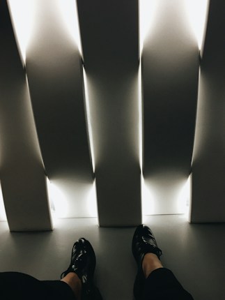The image presents a pair of black shoes worn by an individual seated indoors. The setting includes vertical, white, illuminated panels casting a soft, ambient light across the floor. The overall feel is minimalistic and modern, with strong angular lines and contrasting light and shadow.