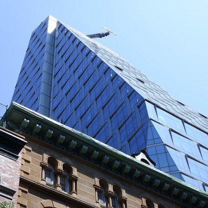 A modern high-rise building with a glass facade is seen towering over an older, ornate brick building. The newer structure features angular architecture and reflective panels, while the older building displays decorative elements and a classic design.