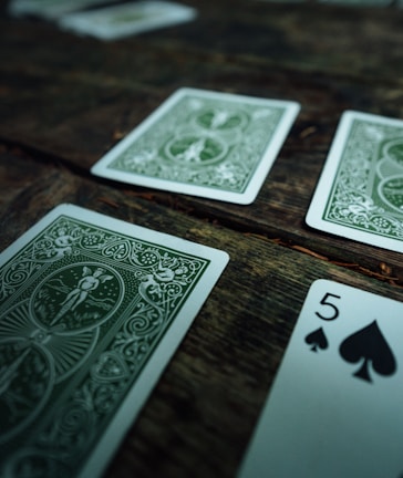 Several playing cards are scattered across a rustic wooden surface. The cards have intricate green designs on their backs, with one card facing up displaying the number five of spades.