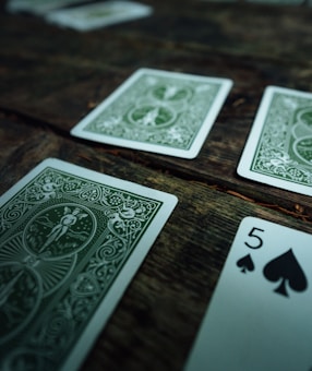 Several playing cards are scattered across a rustic wooden surface. The cards have intricate green designs on their backs, with one card facing up displaying the number five of spades.