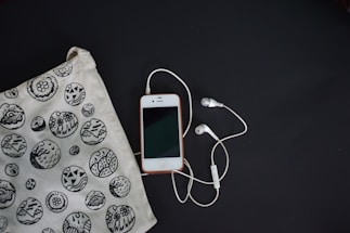 A smartphone with a white front and a connected pair of white earphones is laid on a dark surface. To the left, there is a fabric bag with black and white circular patterns featuring various designs, including abstract and floral motifs.