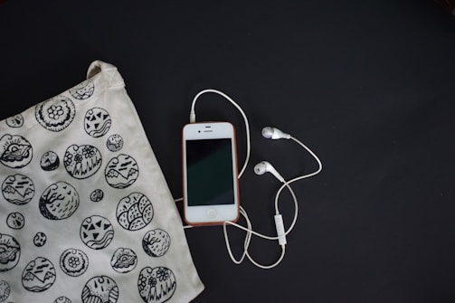 A smartphone with a white front and a connected pair of white earphones is laid on a dark surface. To the left, there is a fabric bag with black and white circular patterns featuring various designs, including abstract and floral motifs.