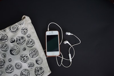 A smartphone with a white front and a connected pair of white earphones is laid on a dark surface. To the left, there is a fabric bag with black and white circular patterns featuring various designs, including abstract and floral motifs.