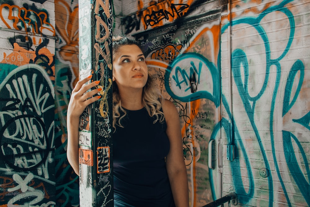 Mujer con camisa negra sin mangas sosteniendo en el poste con graffiti usando la mano izquierda mirando en las paredes con graffiti variado