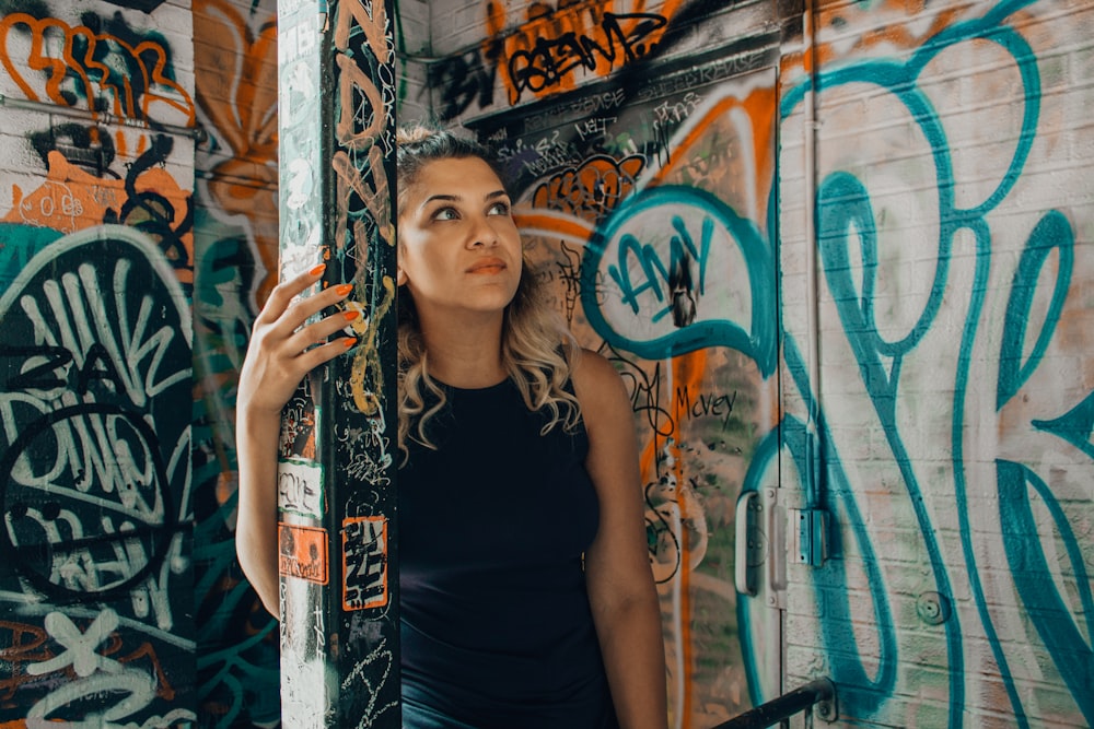 woman wearing black sleeveless shirt holding on post with graffiti using left hand staring on walls with assorted graffiti