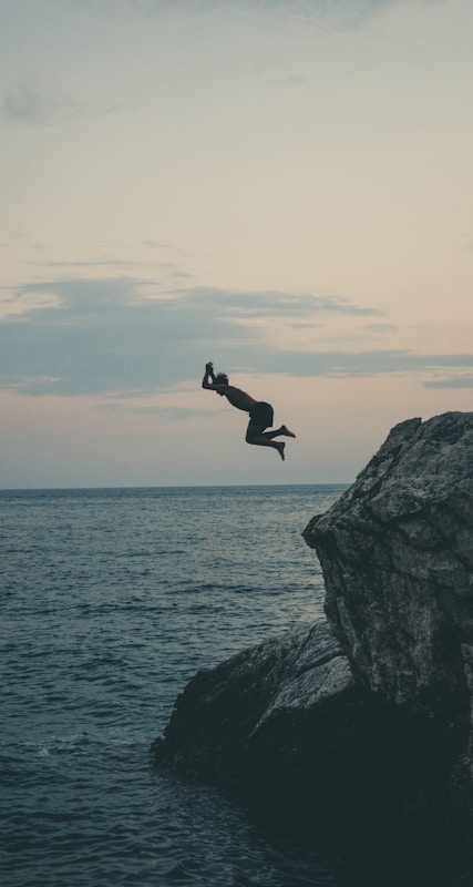 A person is captured in mid-air as they leap off a rocky cliff into the sea below. The sky is overcast with subtle hues indicating dusk or dawn, adding a serene backdrop to the adventurous jump.