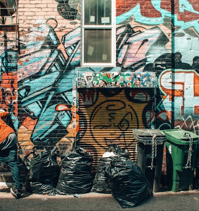 man standing in front of graffiti tagged wall beside trash bags and trash bins at daytime