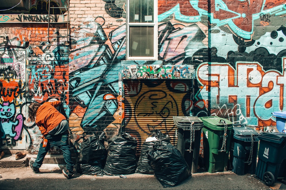 man standing in front of graffiti tagged wall beside trash bags and trash bins at daytime