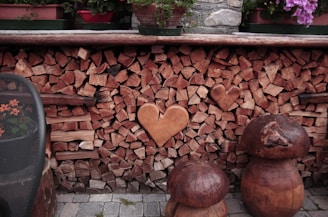 A stack of firewood against a stone wall with heart-shaped wood pieces prominently placed in the center. Wooden stools that resemble mushrooms sit on a stone-paved surface in the foreground. Flower pots with vibrant flowers are placed on top of the firewood stack, adding color to the rustic scene.