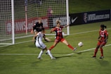 A soccer match with players in action near the goalpost. One player in a white uniform is attempting to control the ball while two players in red and black uniforms are defending. The goalkeeper, also in a dark uniform, is on alert close to the net. The field is well-lit, and banners such as 'NWSL' are visible in the background.