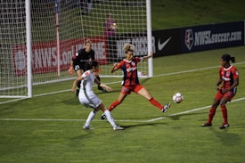 A soccer match with players in action near the goalpost. One player in a white uniform is attempting to control the ball while two players in red and black uniforms are defending. The goalkeeper, also in a dark uniform, is on alert close to the net. The field is well-lit, and banners such as 'NWSL' are visible in the background.