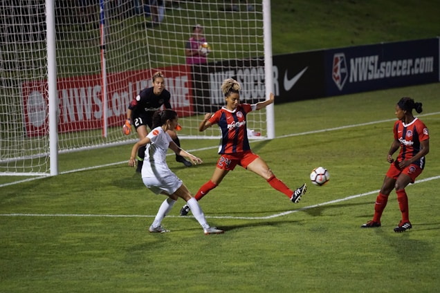 A soccer match with players in action near the goalpost. One player in a white uniform is attempting to control the ball while two players in red and black uniforms are defending. The goalkeeper, also in a dark uniform, is on alert close to the net. The field is well-lit, and banners such as 'NWSL' are visible in the background.