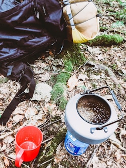 A camping setup in a forested area with a backpack, a rolled sleeping bag, a portable stove with boiling contents, and a red mug placed on the forest floor among autumn leaves and greenery.