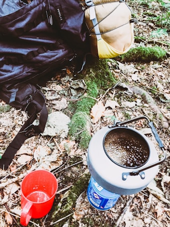 A camping setup in a forested area with a backpack, a rolled sleeping bag, a portable stove with boiling contents, and a red mug placed on the forest floor among autumn leaves and greenery.