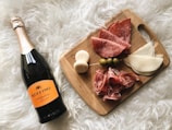 A wooden cutting board displaying a selection of charcuterie including sliced salami, cheese, prosciutto, olives, and small mozzarella balls skewered together. Next to the board is a bottle of Ruffino Prosecco, all set on a fluffy white surface.