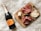 A wooden cutting board displaying a selection of charcuterie including sliced salami, cheese, prosciutto, olives, and small mozzarella balls skewered together. Next to the board is a bottle of Ruffino Prosecco, all set on a fluffy white surface.