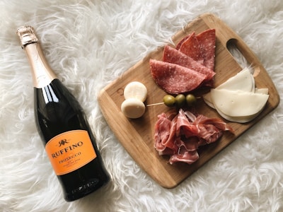A wooden cutting board displaying a selection of charcuterie including sliced salami, cheese, prosciutto, olives, and small mozzarella balls skewered together. Next to the board is a bottle of Ruffino Prosecco, all set on a fluffy white surface.