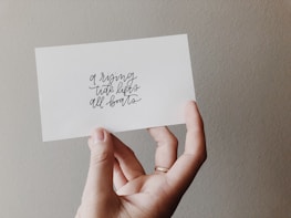 A hand holds a white card with the words 'a rising tide lifts all boats' written in elegant, cursive script. The background features a neutral, textured wall.