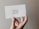 A hand holds a white card with the words 'a rising tide lifts all boats' written in elegant, cursive script. The background features a neutral, textured wall.