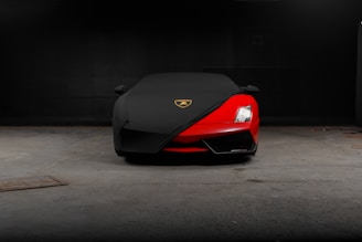 A sleek, red sports car is partially covered with a black protective cover, featuring a gold and black emblem on the cover. The car is parked in a dimly lit garage with a concrete floor.