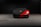 A sleek, red sports car is partially covered with a black protective cover, featuring a gold and black emblem on the cover. The car is parked in a dimly lit garage with a concrete floor.