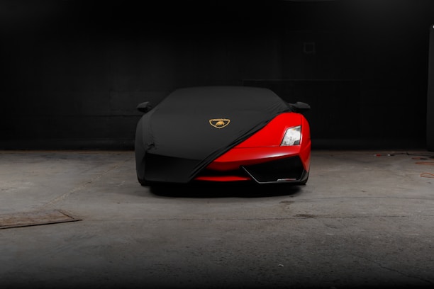A sleek, red sports car is partially covered with a black protective cover, featuring a gold and black emblem on the cover. The car is parked in a dimly lit garage with a concrete floor.