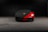 A sleek, red sports car is partially covered with a black protective cover, featuring a gold and black emblem on the cover. The car is parked in a dimly lit garage with a concrete floor.