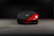A sleek, red sports car is partially covered with a black protective cover, featuring a gold and black emblem on the cover. The car is parked in a dimly lit garage with a concrete floor.