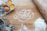 A wooden surface with the word 'Family' written in flour. Surrounding the writing are two brown eggs, one of which is cracked open, a flour sifter, a rolling pin, and a pile of flour. The scene appears to be related to baking.