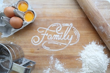 A wooden surface with the word 'Family' written in flour. Surrounding the writing are two brown eggs, one of which is cracked open, a flour sifter, a rolling pin, and a pile of flour. The scene appears to be related to baking.