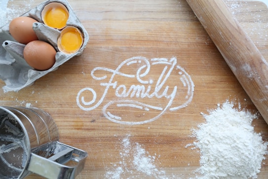 A wooden surface with the word 'Family' written in flour. Surrounding the writing are two brown eggs, one of which is cracked open, a flour sifter, a rolling pin, and a pile of flour. The scene appears to be related to baking.