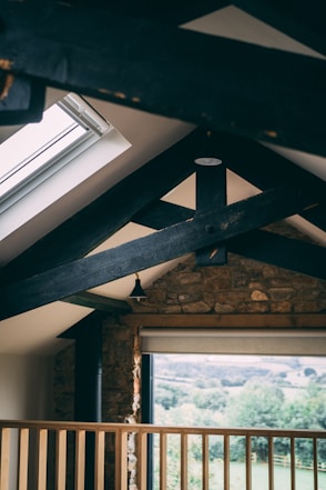 A rustic interior featuring exposed wooden beams and a stone wall. Natural light pours in through a skylight and large window, offering a view of a lush green landscape. A wooden railing adds to the cozy atmosphere.