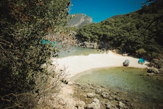 A secluded beach with white pebbles and clear turquoise water is surrounded by lush green hills. Two small boats are beached on the shoreline under the shade of trees, with verdant foliage partially framing the view. The backdrop features a rugged mountain under a clear blue sky.
