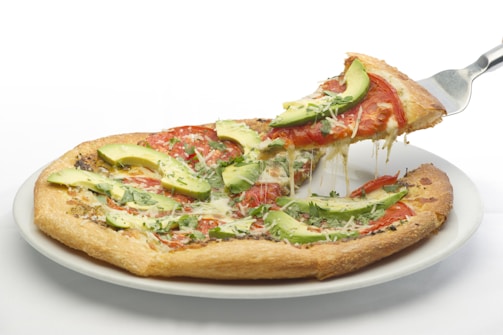 A freshly baked pizza with a slice being lifted, featuring toppings like avocado slices, tomatoes, and a sprinkle of herbs and cheese on a crispy crust.