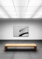 A minimalist gallery space with a geometric design. The room has a large, simple wooden bench placed on a tiled gray floor, facing a modern art piece on the wall depicting a curved staircase. The ceiling features a grid of rectangular lights, casting a neutral ambience.