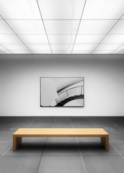 A minimalist gallery space with a geometric design. The room has a large, simple wooden bench placed on a tiled gray floor, facing a modern art piece on the wall depicting a curved staircase. The ceiling features a grid of rectangular lights, casting a neutral ambience.
