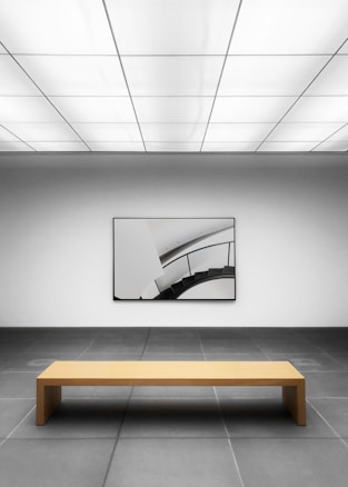 A minimalist gallery space with a geometric design. The room has a large, simple wooden bench placed on a tiled gray floor, facing a modern art piece on the wall depicting a curved staircase. The ceiling features a grid of rectangular lights, casting a neutral ambience.