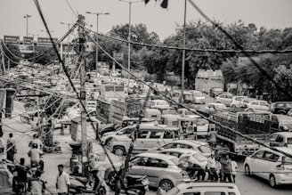 A busy urban street scene with a significant traffic jam, where numerous vehicles including cars, motorcycles, and auto-rickshaws are congested. Overhead, there is a dense network of tangled cables and wires. Pedestrians and vendors can be seen along the sides. The setting suggests a bustling, chaotic environment.