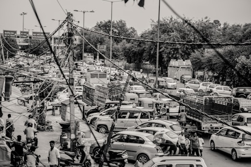 A busy urban street scene with a significant traffic jam, where numerous vehicles including cars, motorcycles, and auto-rickshaws are congested. Overhead, there is a dense network of tangled cables and wires. Pedestrians and vendors can be seen along the sides. The setting suggests a bustling, chaotic environment.