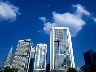 Tall modern skyscrapers with glass facades reach up into a clear blue sky, with some fluffy clouds accentuating the skyline. A large building with the word 'PLATINUM' on its top is prominent in the foreground, while other multi-story buildings stand behind it. The setting suggests an urban cityscape with visible streetlights and a few trees.