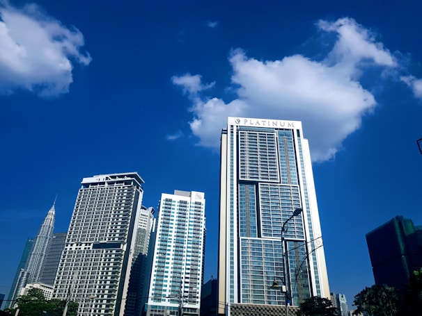 Tall modern skyscrapers with glass facades reach up into a clear blue sky, with some fluffy clouds accentuating the skyline. A large building with the word 'PLATINUM' on its top is prominent in the foreground, while other multi-story buildings stand behind it. The setting suggests an urban cityscape with visible streetlights and a few trees.