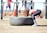 A person in athletic attire appears to be lifting or flipping a large tire in an outdoor gym setting. In the background, other people are seen using gym equipment, suggesting a fitness-focused environment.