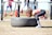 A person in athletic attire appears to be lifting or flipping a large tire in an outdoor gym setting. In the background, other people are seen using gym equipment, suggesting a fitness-focused environment.