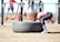 A person in athletic attire appears to be lifting or flipping a large tire in an outdoor gym setting. In the background, other people are seen using gym equipment, suggesting a fitness-focused environment.