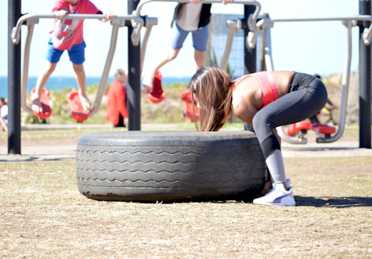 A person in athletic attire appears to be lifting or flipping a large tire in an outdoor gym setting. In the background, other people are seen using gym equipment, suggesting a fitness-focused environment.