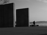 The image depicts a solitary figure walking with a suitcase near two large, monolithic structures by a waterfront. The background shows a calm body of water with distant land and a few visible boats, under a partly cloudy sky. The setting appears peaceful and somewhat isolated.